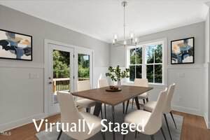 A bright dining room with a wooden table, six white chairs, modern art on the walls, a light fixture above, a window, glass doors to a deck, and the words "Virtually Staged" overlaid on the image.