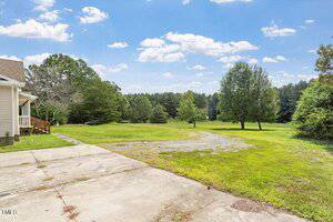 A concrete driveway leads to a grassy yard with scattered trees under a blue sky with white clouds. A house with beige siding is partially visible on the left. Dense wooded areas border the property.