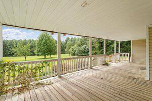 A spacious wooden porch with a white railing overlooks a large grassy yard with several trees. The porch has a white ceiling, and the scene is set on a clear day with blue sky and scattered clouds.