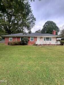 A single-story brick house at 220 Pine Tree Road, Oxford, features a covered front porch with white railing and chimney, set on a large, well-maintained lawn with trees in the background under a cloudy sky.