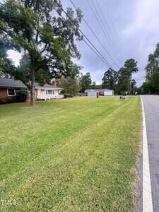A large, neatly mowed lawn stretches alongside a paved road, bordered by a brick house with a porch on the left and a white detached garage in the background. Overhead power lines run along the street.