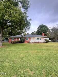 A single-story red brick house with white trim and a front porch sits on a large, neatly mowed lawn. Mature trees surround the property, and the sky appears overcast.