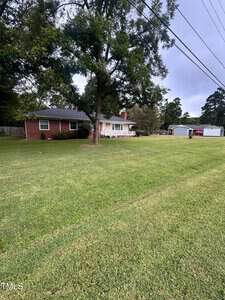 A single-story brick house with white trim sits behind a large, well-kept lawn. There are several trees, a driveway, a detached garage, and power lines overhead under a cloudy sky.