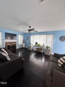 A living room with light blue walls, dark wood floors, two dark sofas, a fireplace with a TV above it, a ceiling fan, and a window with two desks underneath holding houseplants.