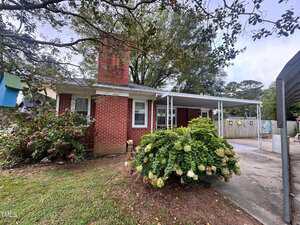 Single-story red brick house with a prominent chimney, white trim, and a carport on the right. Large bush with flowers in front, tree branches above, and paved driveway visible. Fence and yard in the background.