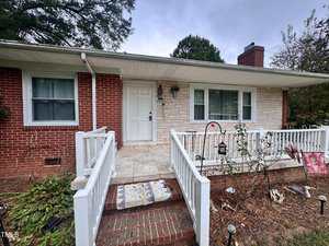 A single-story brick and stone house with a white front door, large front windows, and a white railing along a small porch. The front yard has scattered plants and a few items, with a paved walkway leading to the entrance.