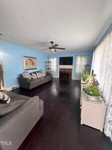 A living room with light blue walls, dark wood floors, two gray couches, a ceiling fan, a fireplace with a TV above it, and large windows with patterned curtains. There are plants on a white cabinet.