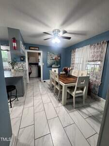 A dining area with a wooden table and six white chairs, a ceiling fan with light, blue walls, large tile floor, floral curtains, a wall painting, and a kitchen area visible to the left.