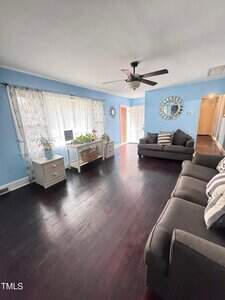 A living room with light blue walls, dark wood flooring, two gray sofas, white side tables, a ceiling fan, large window with sheer curtains, a wall mirror, and several potted plants near the window.