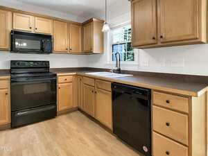 A kitchen with light wood cabinets, black appliances including a microwave, stove, and dishwasher, a white sink below a window, and light wood flooring. The countertops are dark brown.