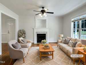 A bright living room with a beige sofa, two armchairs, a wooden coffee table, a fireplace, and a ceiling fan. Large windows let in natural light. The image is marked as "virtually staged.