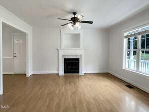 A bright, empty living room with light wood flooring, a white fireplace centered on one wall, a ceiling fan with lights, large windows on the right, and a white door to the left.