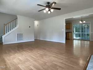 A spacious, empty living room with light wood flooring, white walls, a ceiling fan with lights, a staircase to the left, and an open view into the kitchen and dining area with large windows.