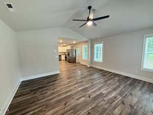 A spacious, empty living room with wood-look flooring, white walls, and a ceiling fan. The room opens to a kitchen with stainless steel appliances and several windows letting in natural light.