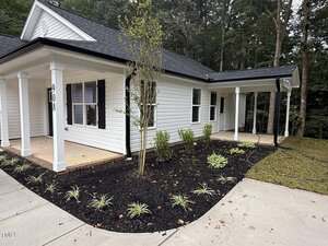 A white single-story house with black shutters and a covered porch is shown. The front yard features fresh landscaping with small shrubs, mulch, and young trees. A driveway and sidewalk run alongside the house.