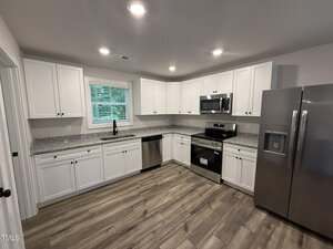 Modern kitchen with white cabinets, stainless steel appliances, granite countertops, a dishwasher, and wood-look flooring. A large window above the sink brings in natural light, and recessed lighting brightens the room.