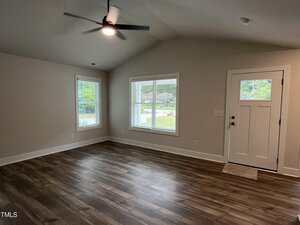 A room with wood-look flooring, beige walls, two windows with white blinds, a white front door with glass panels, and a ceiling fan with a light. The room is empty and well-lit with natural light.