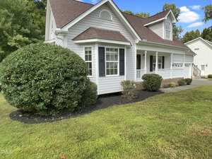 A white single-story house with a covered front porch, dark shutters, and a gable roof. There are trimmed bushes and green grass in the yard under a blue sky with trees in the background.