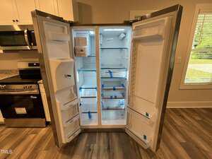 A new, empty side-by-side refrigerator with both doors open, showing clean shelves, drawers, and storage compartments. Blue tape secures the shelves. The kitchen floor, cabinets, and a window are visible.