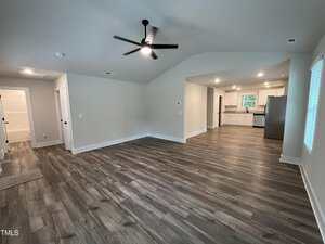 Spacious open-concept living area with wood-look flooring, white walls, and vaulted ceiling. The room connects to a modern kitchen with white cabinets, stainless steel appliances, and a window above the sink.