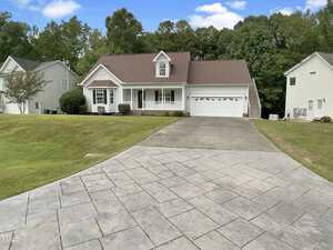 A single-story house with a front porch, attached two-car garage, and sloped driveway. The home has white siding and is surrounded by a lawn and neighboring houses, with trees in the background.