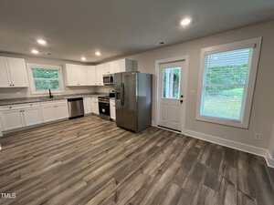 A modern kitchen with white cabinets, stainless steel appliances, wood-look flooring, two windows, and a glass-paneled door leading outside. The walls are light-colored, and the space appears clean and uncluttered.