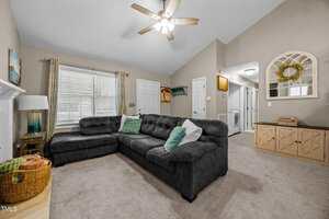 A living room with a large dark gray sectional sofa, light carpeted floor, ceiling fan with light, window with curtains, wall art, and a hallway leading to other rooms. Cabinet and laundry area visible in the background.