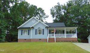 A single-story house with light blue siding, white trim, and a brick foundation, featuring a front porch with railing, an American flag, a concrete driveway, and a lawn bordered by tall trees.