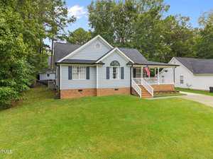 Single-story house with gray siding, white trim, and brick foundation. It has a front porch with white railings, an American flag, a sloped driveway, and a well-kept lawn. Trees and neighboring houses are in the background.
