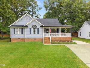 A single-story house on 2771 Clifton Avenue in Creedmoor with gray siding, white trim, dark shutters, a brick foundation, front porch with railing, American flag, and driveway, surrounded by green lawn and trees.