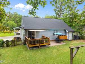 A small house with gray siding and a dark roof features a wooden deck with a sunshade, a patio area with an umbrella and grill, a fenced backyard, and green grass surrounded by trees.
