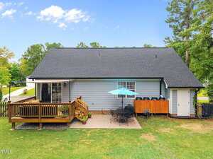 A backyard view of a house with gray siding, a wooden deck with stairs, a patio area with a table, chairs, and blue umbrella, and a fenced grassy yard bordered by trees and a street in the background.