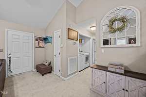 A laundry area with a washer and dryer is visible through a doorway. The wall is beige, and there is a console table with a mirror and wreath above, a bench, and framed artwork on the walls. Carpeted floor throughout.