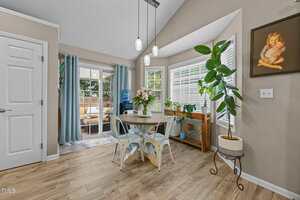 A small dining area with a round wooden table, four chairs, a potted plant, and flowers. Large windows and glass doors let in natural light. Pendant lights hang overhead, and a framed artwork is on the beige wall.