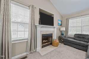 A living room features a lit fireplace with a white mantel, a flat-screen TV above it, two windows with curtains, a gray sectional sofa, a floor lamp, and a basket with blankets on the carpeted floor.