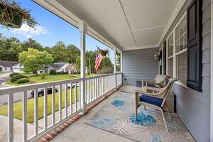 A covered front porch with two wicker chairs, a patterned rug, white railing, and hanging plants overlooks a suburban street with houses, trees, and an American flag on display.