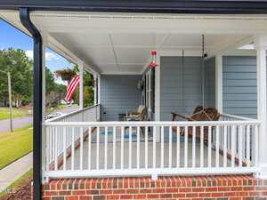 A covered front porch with white railing, brick steps, a wooden swing, two chairs, a small table, a red bird feeder hanging from the ceiling, and an American flag on the left side. A quiet street and trees are visible.