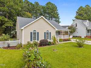 Single-story beige house with a gray roof, white trim, and a covered front porch. Well-maintained lawn, shrubs, and flowers in the front yard. White picket fence and neighboring house visible. Tall trees in the background.