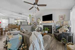 A living room with a ceiling fan, a large sectional sofa, plants, a fireplace with a black TV above it, and various framed photos and decorations on the walls. A dining area is visible in the background.