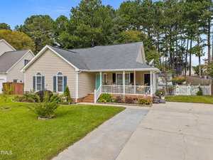 One-story beige house with a gabled roof, white trim, small front porch, and attached garage. Well-maintained yard with shrubs and a curved driveway. Tall pine trees and a white fence are visible in the background.