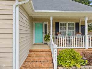 Front porch of a house with beige siding, brick steps, a turquoise front door, white railing, two rocking chairs, potted plants, and the house number 2535 visible on a white post.