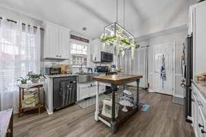 A bright kitchen with white cabinets, stainless steel appliances, a farmhouse sink, and a wooden island. The island has shelves underneath and a hanging light fixture with greenery above. Light wood floors and sheer curtains complete the space.