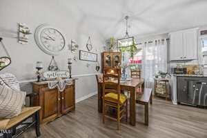 A dining area with a wooden table and four chairs, a wooden side cabinet, wall decorations including a large clock and "FAMILY" sign, light wood flooring, and bright natural light from a window.