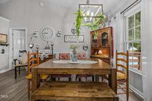 A dining room with a wooden table, bench, and chairs. A cabinet with dishes, a wall clock, and decor including a "FAMILY" sign are visible. Plants hang from a light fixture above the table. Large window with white curtains.