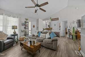 A living room with a gray sofa, blue armchair, wooden coffee table, and ceiling fan. Light pours in through large windows with white curtains. Decorative items and plants are placed around the room.