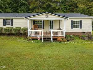 A single-story beige house with a metal roof, black shutters, and a covered porch with a hanging chair. The front yard is grassy with some shrubs and flower beds along the house. Trees are visible in the background.