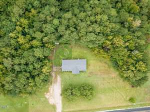 Aerial view of a single-story house with a gray roof, surrounded by trees on three sides and a large grassy yard, adjacent to a paved road.