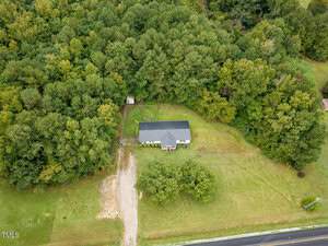Aerial view of a single-story house with a dark roof, surrounded by dense trees and a large grassy yard. A driveway leads from the road to the house, with some cleared patches visible near the entrance.