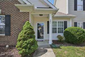 A front entrance of a house with a glass storm door, brick and siding exterior, windows with black shutters, two bushes, and a concrete walkway leading to the door, beneath a small covered porch.