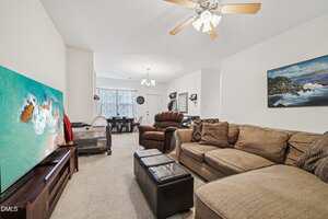 A living room with a beige sectional sofa, brown recliner, and ottoman, facing a large TV displaying an aerial beach scene. The space extends to a dining area with a table and chairs near a window.